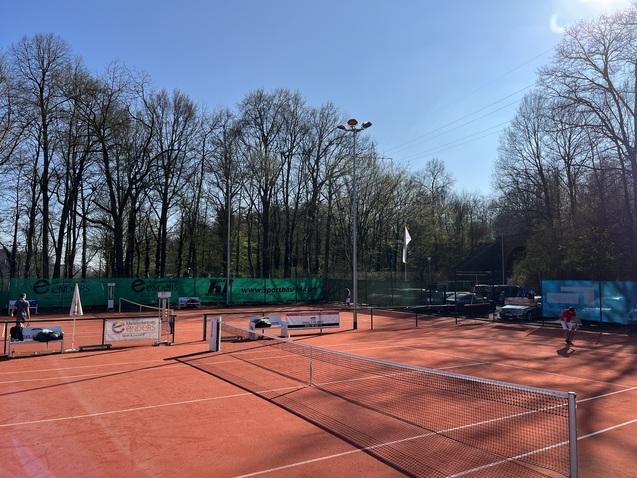 Tennisplatz mit rotem Sand, Bäumen im Hintergrund und Spielern bei sonnigem Wetter.