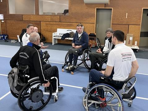 Sechs Rollstuhlfahrer sitzen in einem Sportzentrum und diskutieren auf einer blauen Sportfläche.