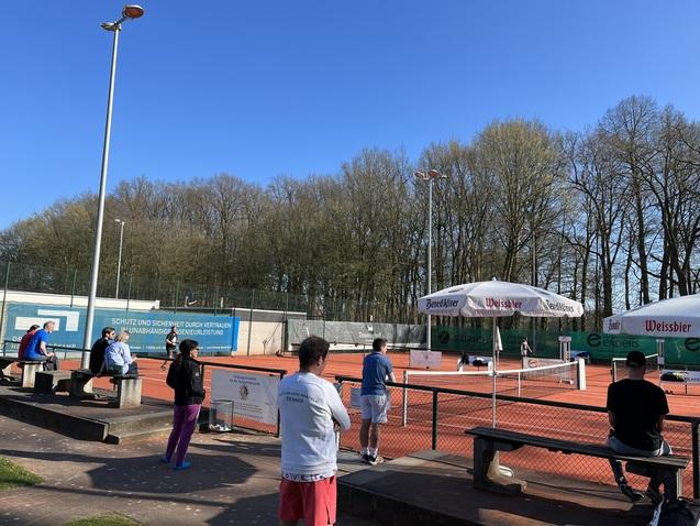 Gruppierung von Personen auf Bänken an einem Tennisplatz, im Hintergrund Tennis-Spieler unter blauem Himmel.
