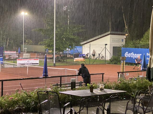 Regnerische Szenerie mit einem Tennisspieler auf einem roten Platz, Blick von einem überdachten Sitzbereich.