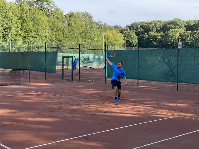 Tennisspieler in blauem Shirt schlägt einen Ball auf einem Sandplatz mit grünen Umzäunungen und Bäumen im Hintergrund.