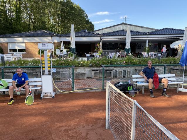Zwei Männer sitzen auf Bänken neben einem Tennisplatz, der Hintergrund zeigt ein Clubhaus und Bäume.