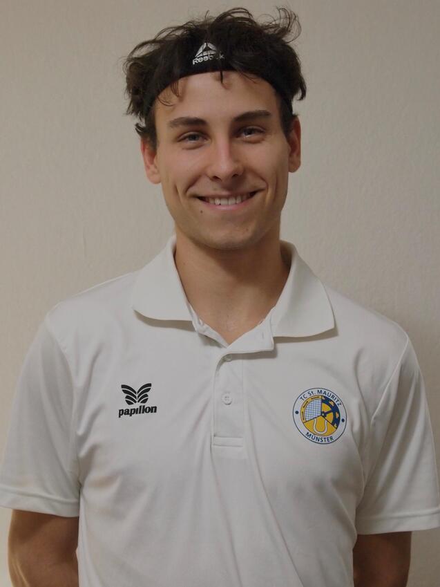 Lächelnder junger Mann in weißem Poloshirt mit Vereinslogo und Kopfband vor neutralem Hintergrund.