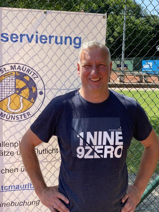 Mann mit kurzem Haar steht vor einem Tennisplatz und einem Schild zur Platzreservierung in Münster.