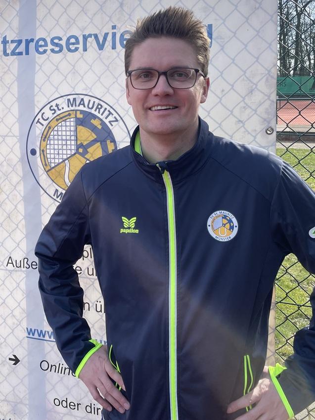 Mann mit Brille und Sportjacke steht lächelnd vor einem Banner mit dem Logo des TC St. Mauritz und Platzreservierungsinformationen.
