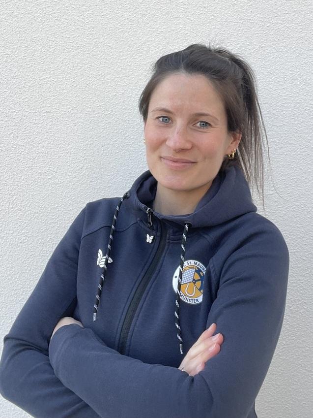 Porträt einer lächelnden Frau mit schulterlangen Haaren, die eng verschränkte Arme vor einem neutralen Hintergrund hält.