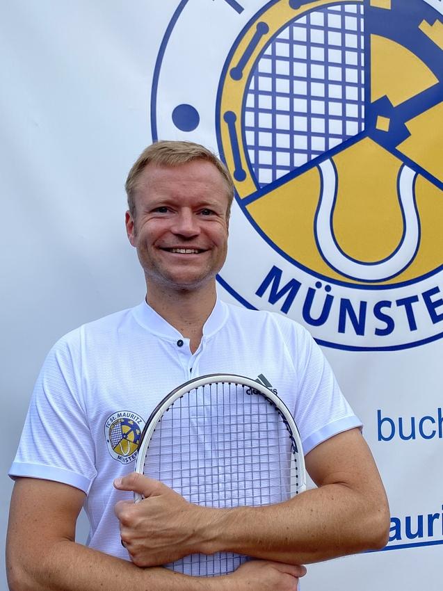 Lächelnder Mann in weißem Shirt hält einen Tennisschläger vor einem Banner des TC St. Mauritz Münster.