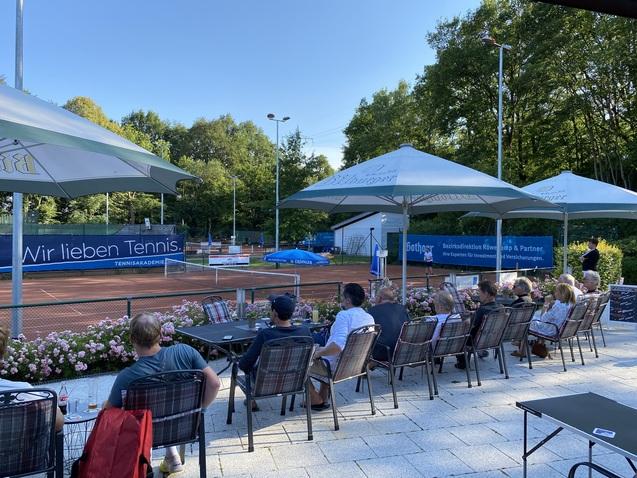 Blick auf einen Tennisplatz mit Zuschauern in Stühlen unter Schirmen, umgeben von Blumenbeeten und Bäumen.