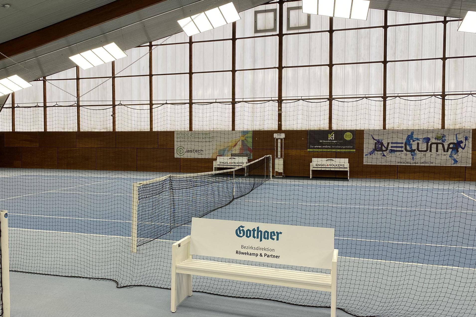 Innenansicht einer Tennishalle mit blauem Boden, Netzen und Bänken an den Seiten. Großer Fensterbereich im Hintergrund.