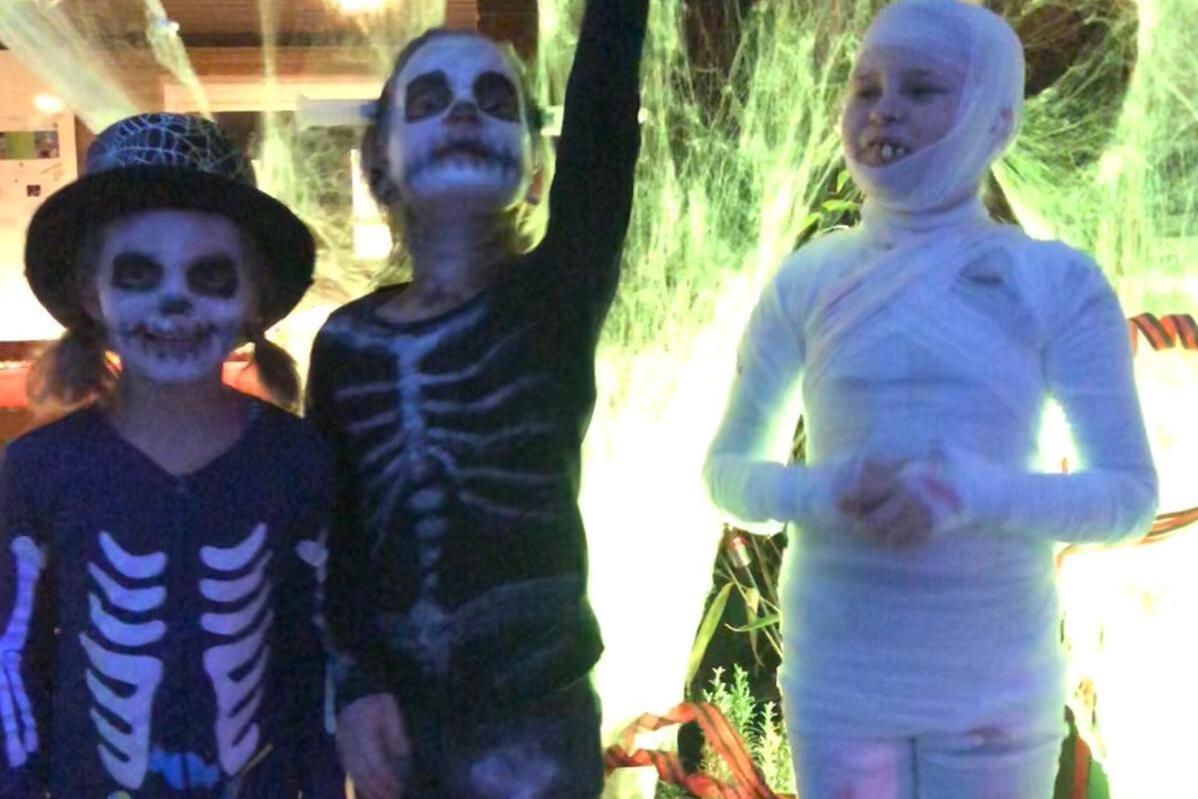 Drei Kinder in Halloween-Kostümen: zwei als Skelette, eines als Mumie, vor einer gruseligen Dekoration aus Spinnweben.