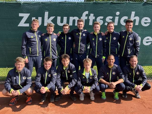 Gruppe von 14 jungen Tennis-Spielern in Sportjacken, lächelnd vor einem grünen Netz auf einem Tennisplatz.