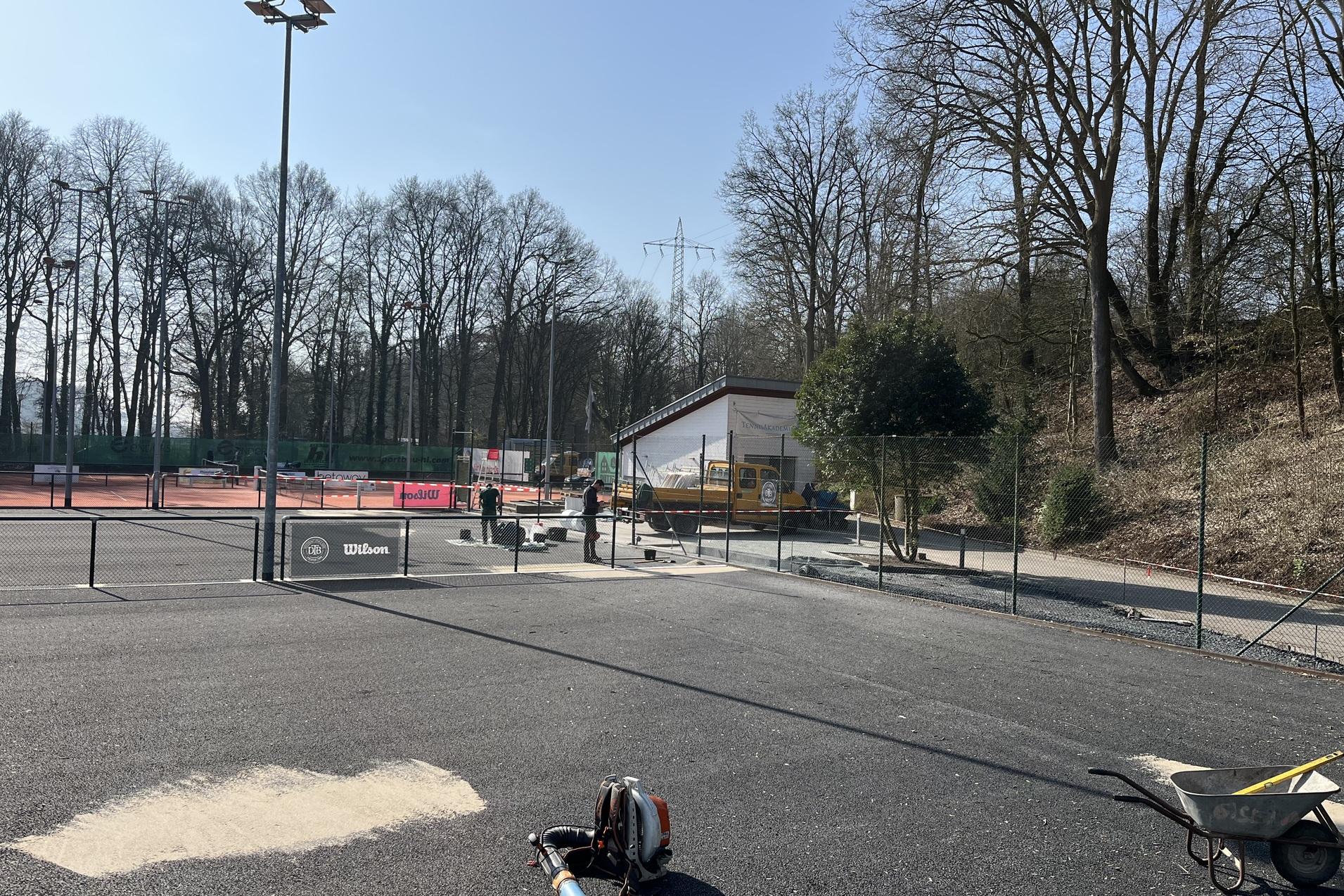 Blick auf einen Tennisplatz mit Bauarbeiten, einer Baumaschine und umgebenden Bäumen unter blauem Himmel.