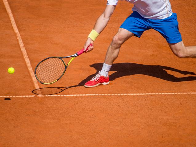 Ein Tennisspieler schlägt mit einem Racket einen gelben Ball auf einem roten Sandplatz.