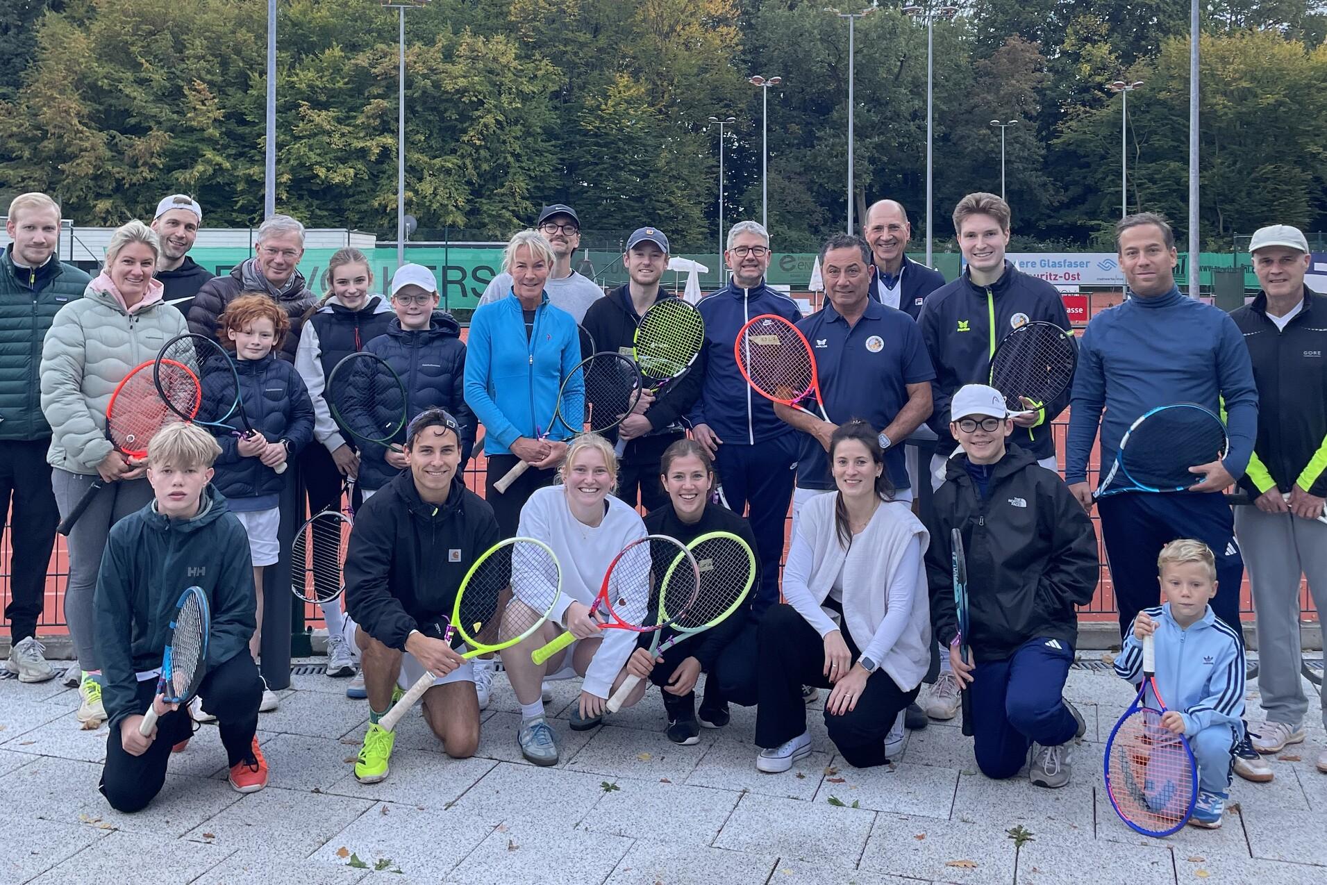 Gruppenshot von 24 Personen auf einem Tennisplatz, alle halten Tennisschläger, mit Bäumen im Hintergrund.