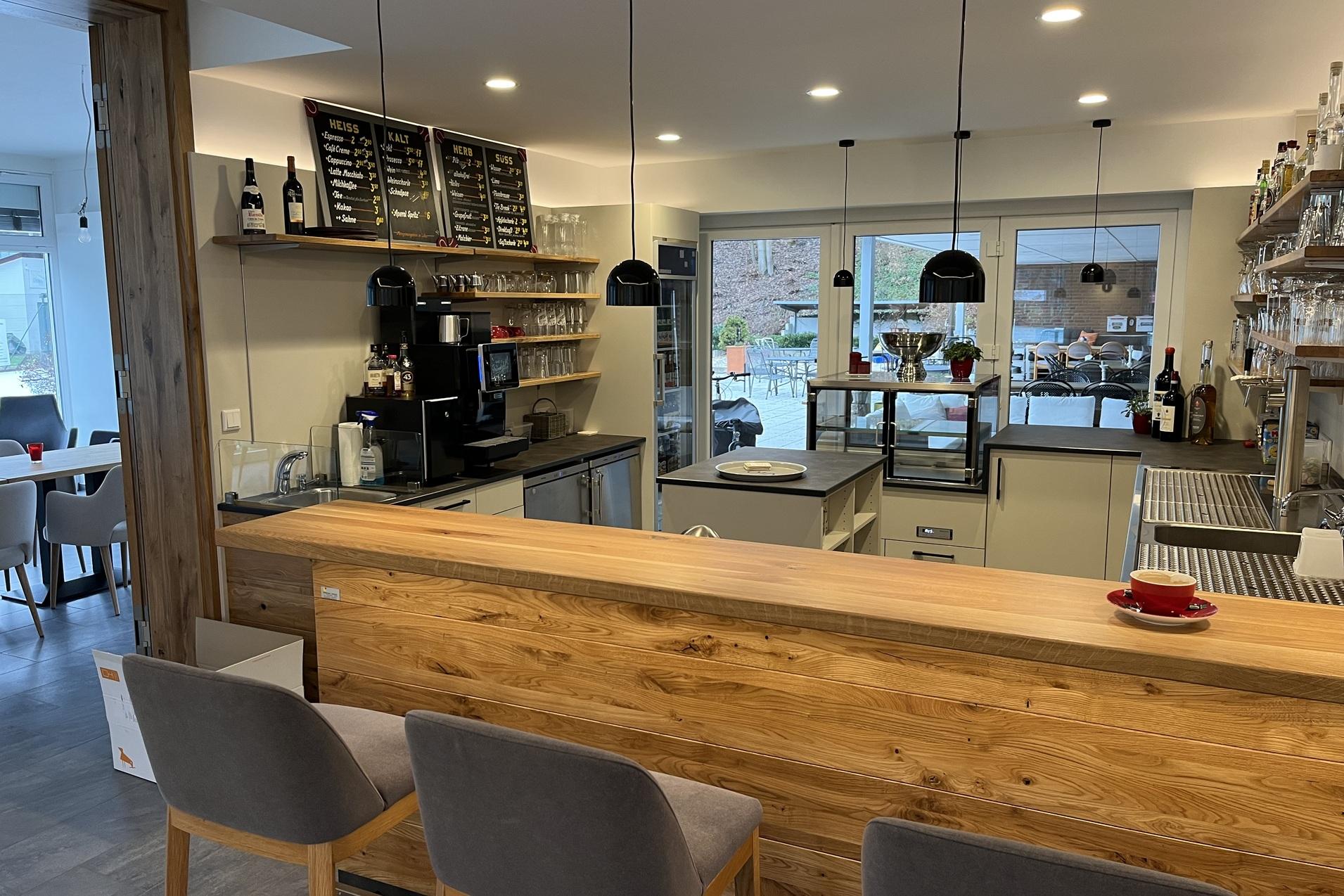 Moderne Cafébar mit Holztheke, grauen Stühlen und offenem Küchenbereich. Tageslicht fällt durch große Fenster.